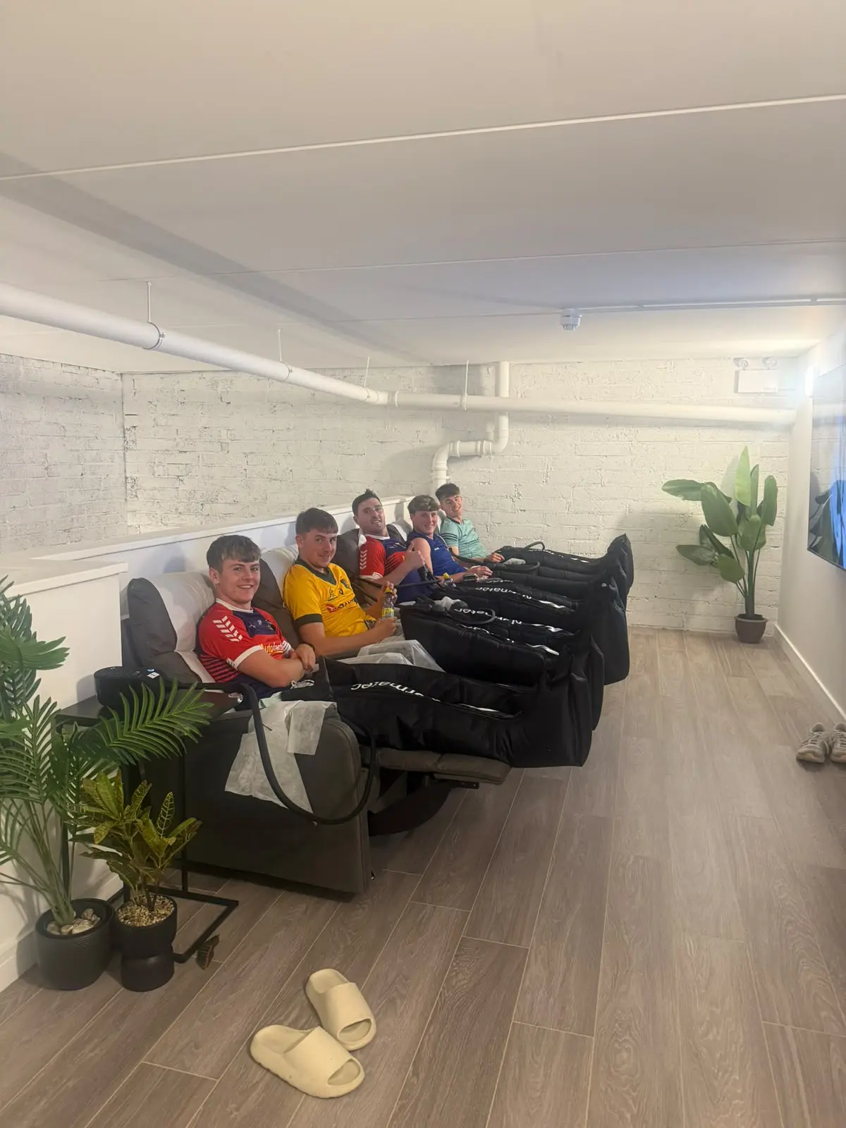 A group in team jerseys reclining with the compression boots on, slippers on the wooden floor